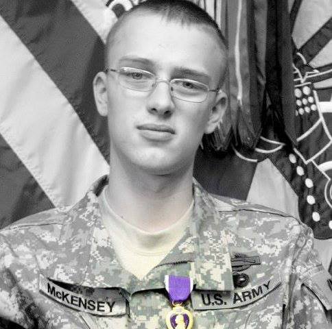 Black and white image of young soldier with glasses in camouflage, with Purple Heart pin on collar